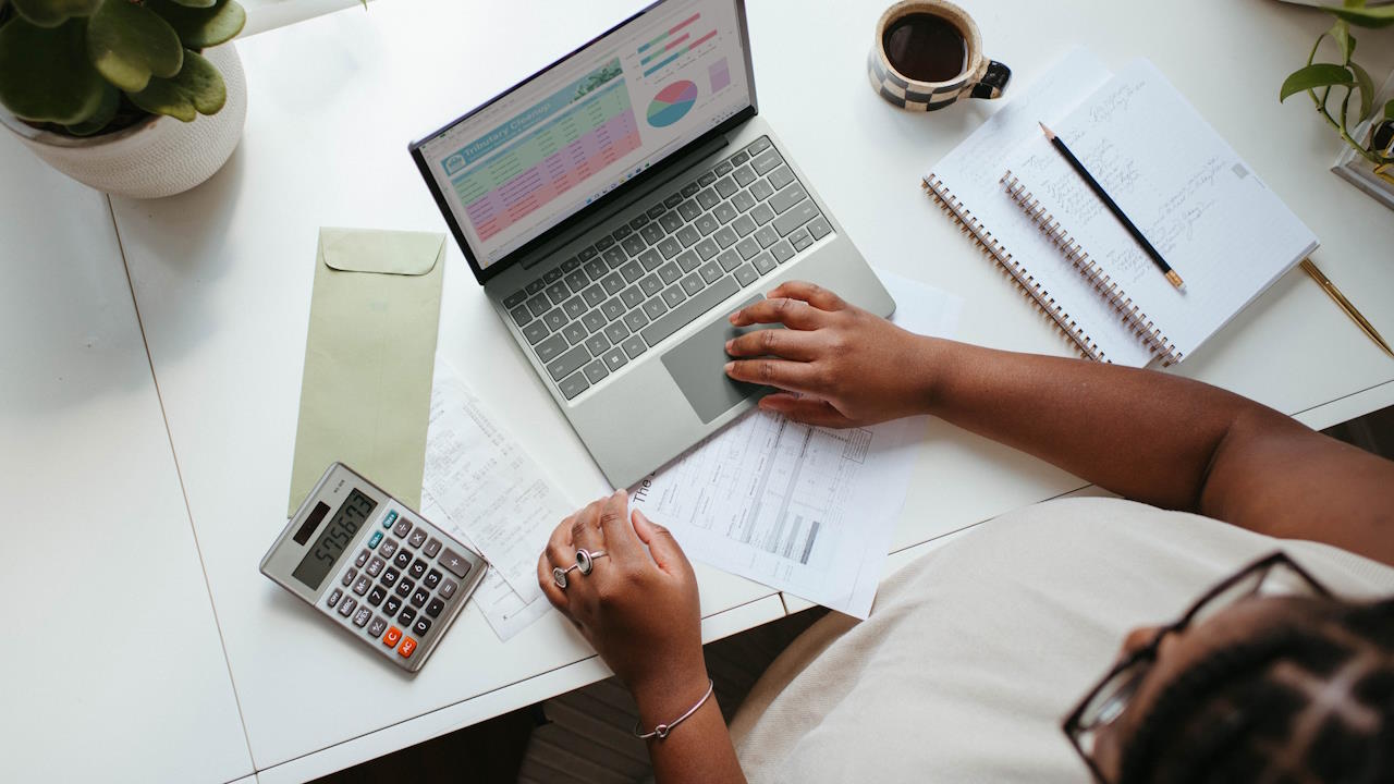 A young woman sitting at a table with a laptop, several notebooks, and a calculator