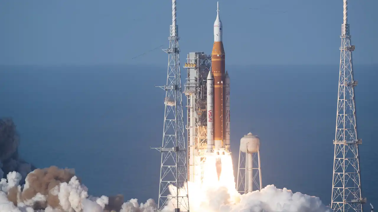 A wide shot of the Artemis SLS rocket launching from Kennedy Space Center. The orange core stage and twin white solid rocket boosters emit a bright white glare and thick clouds of exhaust. Three tall metal lightning towers frame the launch pad against a clear, pale blue horizon.