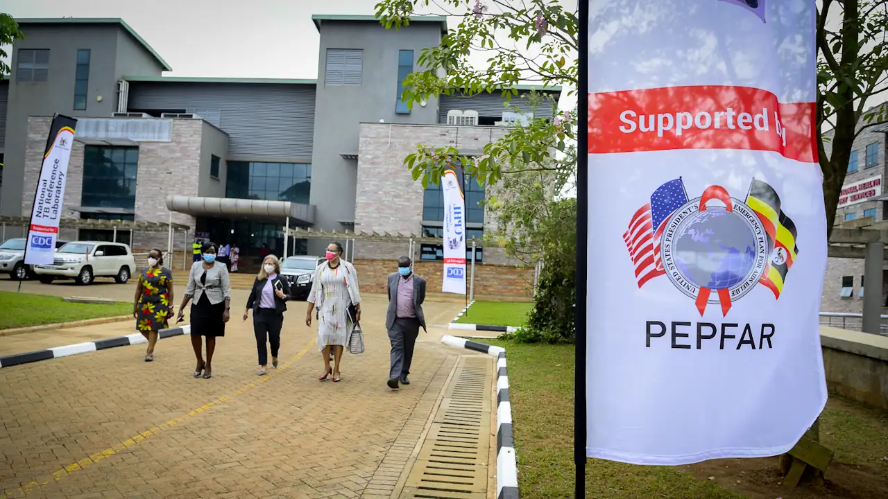Exterior shot of the CDC-supported National Health Laboratory and Diagnostic Services facility in Kampala, Uganda, 2022. U.S. Ambassador Natalie A. Brown and others walk towards the entrance. A PEPFAR flag is prominently displayed.