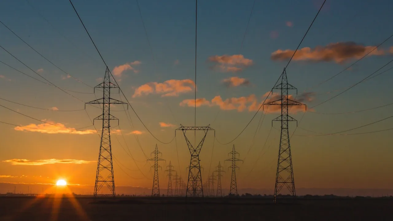 A row of steel lattice electricity pylons carrying power lines across a rural landscape during dawn.