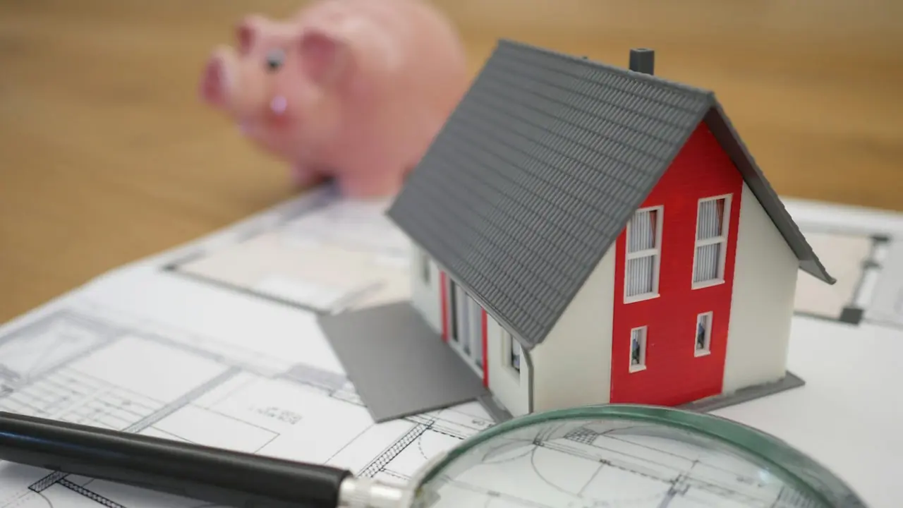 An illustration for property, mortgages, bank loans and home savings. A model of a house sitting on architectural plans, with a blurred piggy bank in the background.