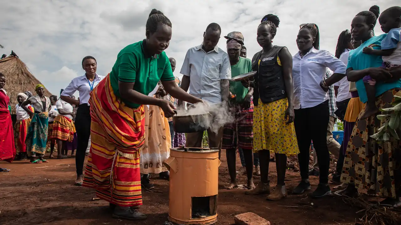 A group of people, mostly women, watch a woman place a steaming saucepan on a stove.