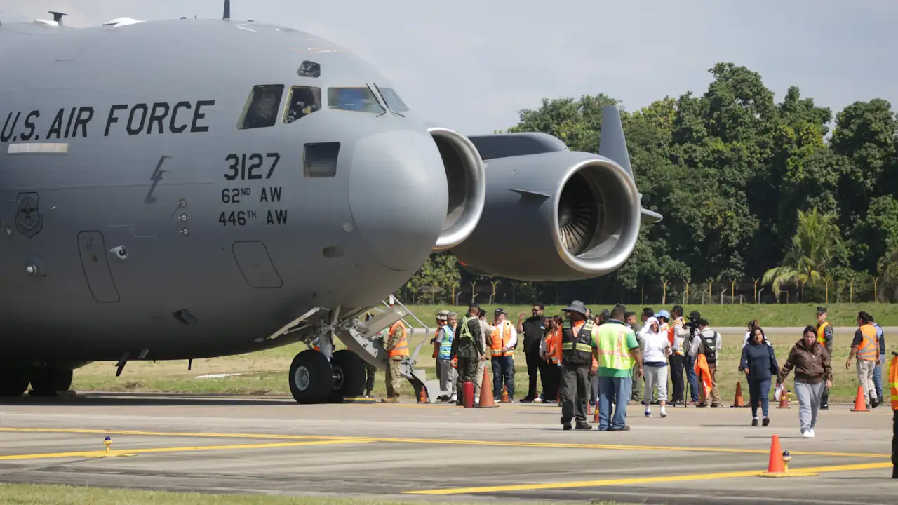 Close-up profile of the nose and engines of a US Air Force C-17 Globemaster III aircraft on a sunny day. Markings on the fuselage identify the 62nd and 446th Airlift Wings. On the tarmac below, a group of civilians and officials in high-visibility vests stand near the aircraft steps.