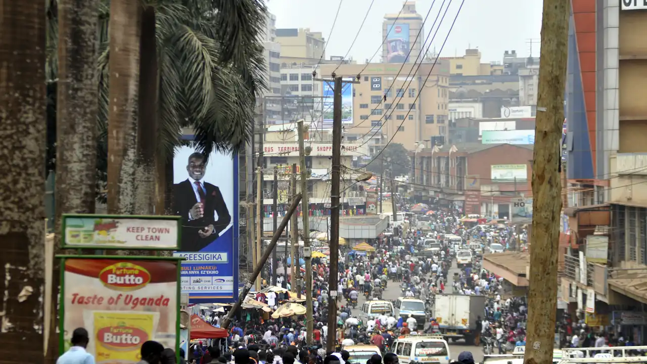 A high-angle view of a crowded Namirembe Road in downtown Kampala, featuring a dense flow of pedestrians, commuters, and vehicles amidst commercial buildings and advertising billboards.
