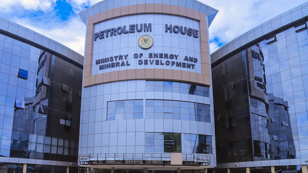 A low-angle view of a modern, glass-fronted office building against a partly cloudy blue sky. The central facade bears the words "Petroleum House" above "Ministry of Energy and Mineral Development," separated by a circular government emblem.