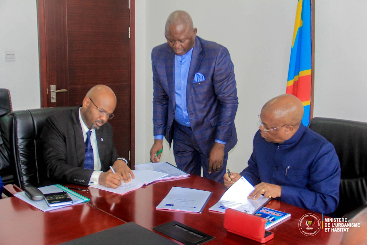 Two men, seated, sign an agreement in an office, with an aide to one of them standing between them