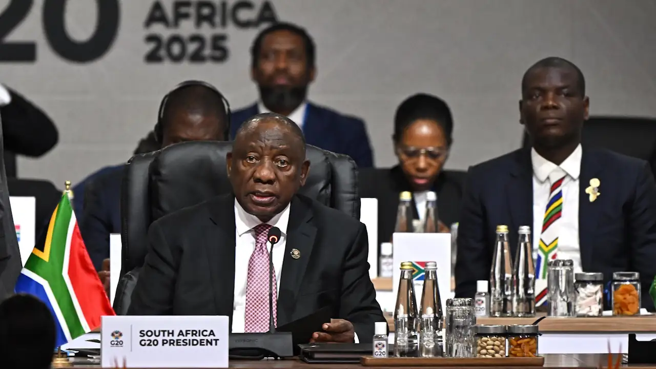 South African President Cyril Ramaphosa sits at a desk with a placard reading "South Africa G20 President," backed by a "South Africa 2025" banner and delegation members.