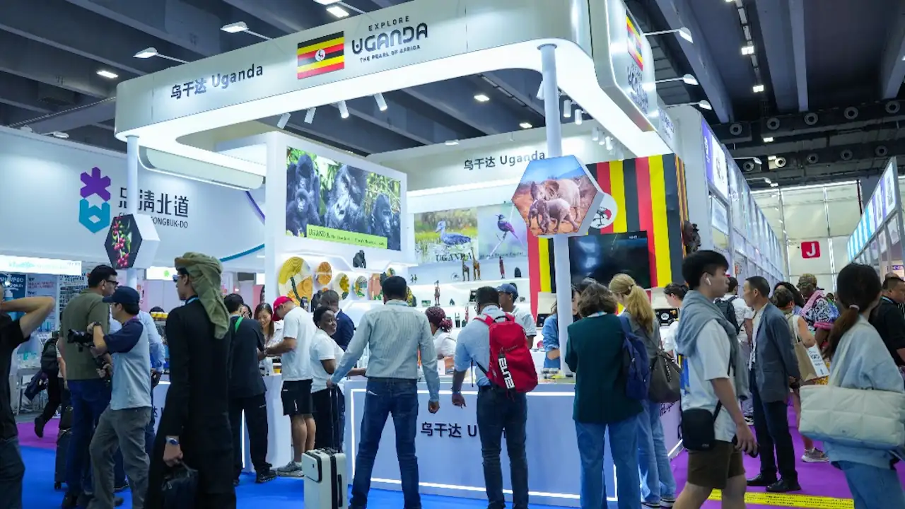 A brightly lit, modern exhibition stall for Uganda at the 138th Canton Fair. The stall features the national flag and the "Explore Uganda" branding, alongside large posters of gorillas and elephants. A diverse crowd of business professionals and visitors are gathered around the counters, discussing trade opportunities in agriculture, coffee, and tourism.