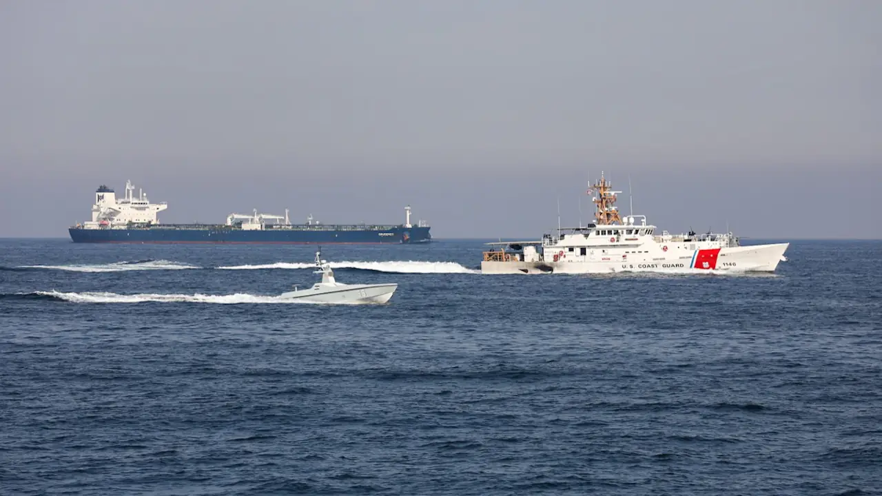 A US Coast Guard cutter sails alongside a sleek, smaller unmanned surface vessel in open blue water, with a large commercial tanker visible on the horizon.