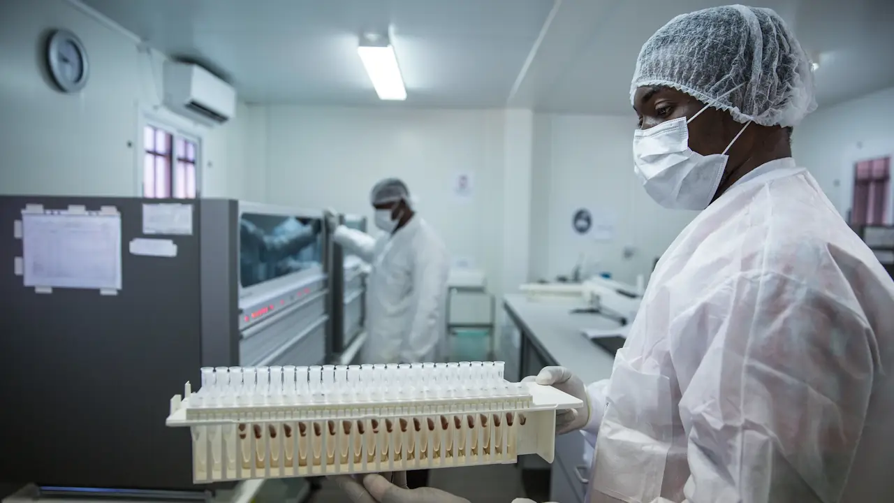 A laboratory technician in white protective gear and a face mask holds a rack of test tubes in a bright, modern molecular biology lab in Mozambique. In the background, another technician works at a large diagnostic machine.